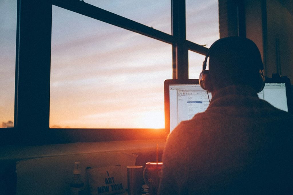 Person sitting behind a laptop with headphones on - sunset visible in a large window in the background
