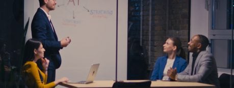 A man in a navy suit stands at a whiteboard presenting to three seated colleagues in a meeting room, with “AI” circled at the centre of a diagram linking plan, goals, strategy and content